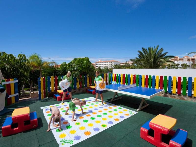 Bunter Spielplatz mit Rutschen, Seilnetz, großer Matte und Sitzblöcken unter blauem Himmel.