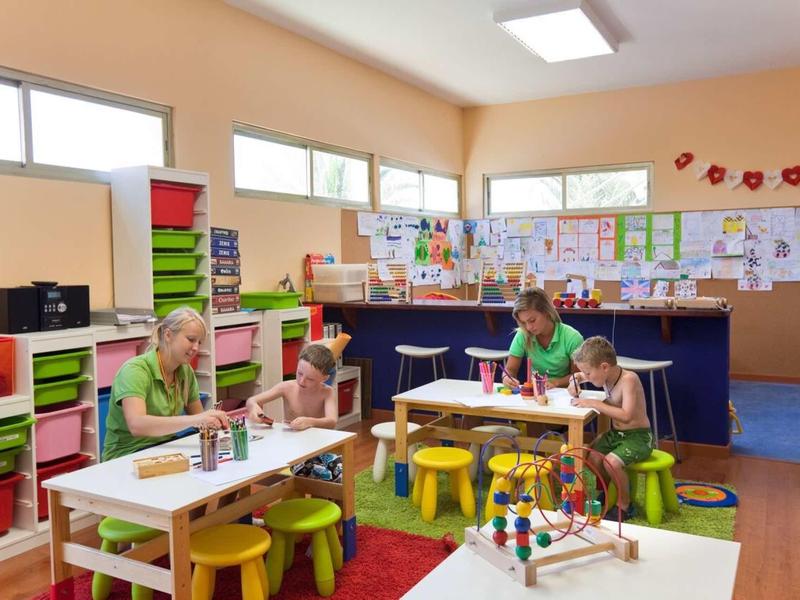 Klassenzimmer mit Kinderbänken, bunten Stühlen, Lehrerin und Kindern beim Malen.