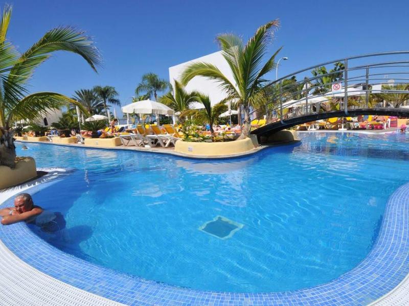 Großer blauer Pool mit Palmen, Brücke und entspannten Menschen bei sonnigem Himmel.