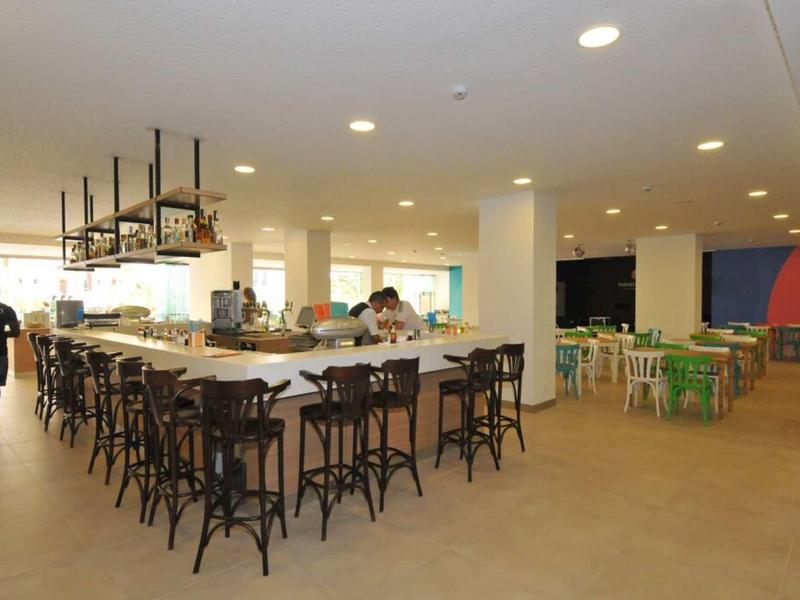 Großer, heller Raum mit langer Bar, Holzstühlen und Sitzgruppen aus bunten Stühlen und Tischen.