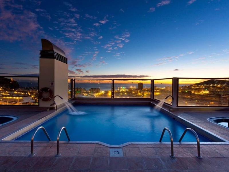Schwimmbecken auf Dachterrasse bei Sonnenuntergang mit Glasgeländer und Stadtblick im Hintergrund.