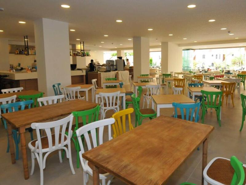 Großer hell erleuchteter Speisesaal mit Holztischen und bunten Stühlen aus Kunststoff.