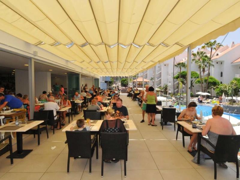 Große überdachte Terrasse mit vielen Tischen, Menschen beim Essen neben einem Pool und Gebäuden.