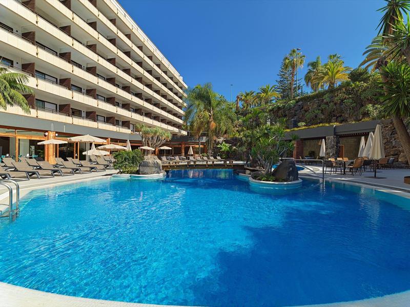 Piscina blu circondata da un hotel moderno e palme sotto il sole.
