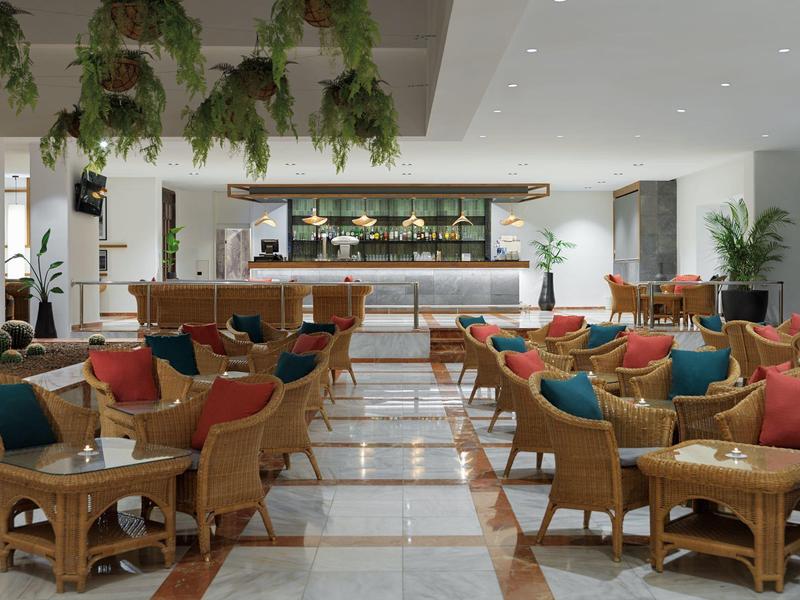 Ampia area salotto di un hotel con poltrone in legno e cuscini colorati.