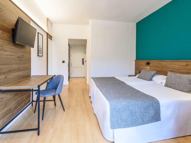 Camera d'hotel moderna con letto matrimoniale, scrivania, sedia blu e TV a muro.