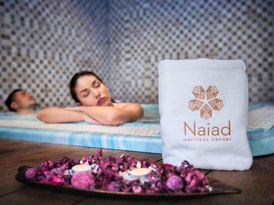 Zwei Personen entspannen in einem Spa, daneben ein Handtuch mit dem Schriftzug "Naiad" und Blumen.