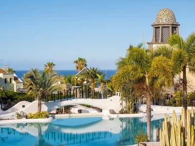 Blick auf einen Pool mit Brücke, Palmen und einem Turm vor klarem blauem Himmel und Meer.