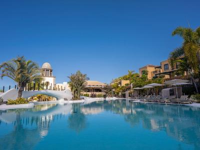 Großer blauer Pool vor weißen Gebäuden mit Palmen unter klarem blauem Himmel.