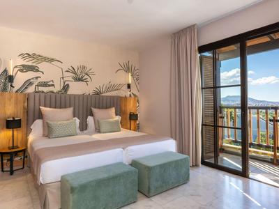 Helles Schlafzimmer mit Doppelbett, grünen Sitzbänken, Pflanzenmotiven und Balkon mit Meerblick.