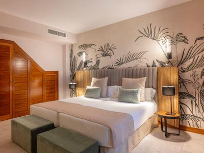 Helles Schlafzimmer mit großem Bett, gemusterter Wand, Holzschrank und zwei grünen Hockern.