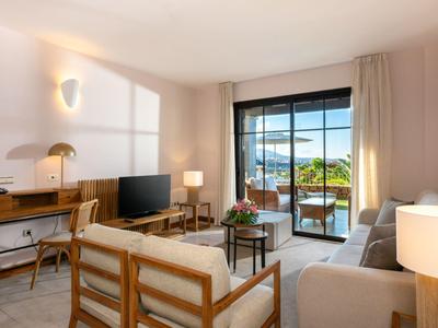 Helles Wohnzimmer mit Holz- und Beiger Möbeln, großem Fenster und Balkon mit Blick ins Grüne.