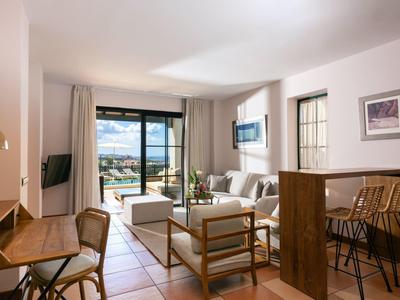 Helles Wohnzimmer mit Sofa, Holzstühlen, Schreibtisch und großen Fenstern mit Meerblick.