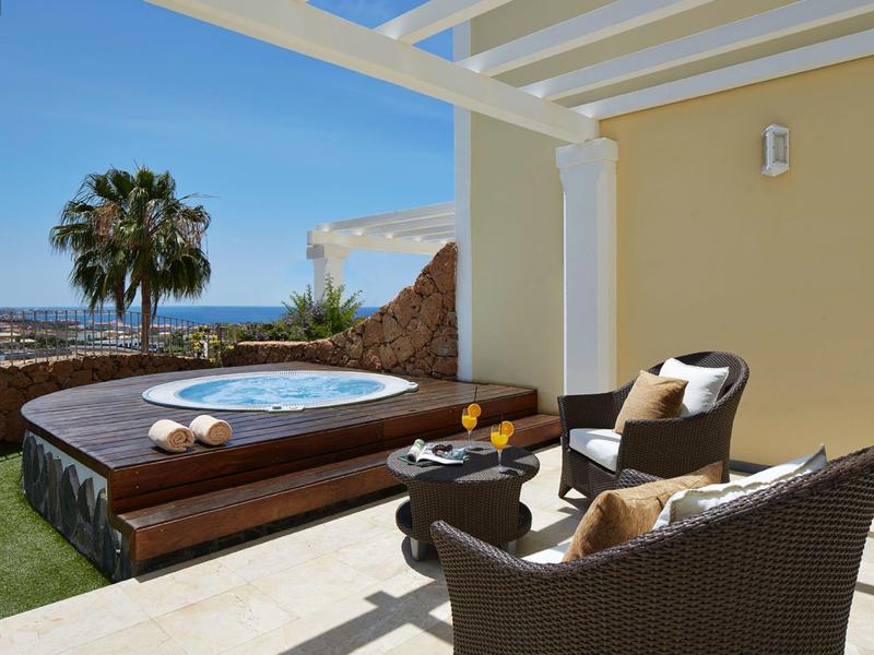 Terrasse mit Whirlpool, zwei Korbsesseln, Tisch, Meerblick und Palmen unter blauem Himmel.