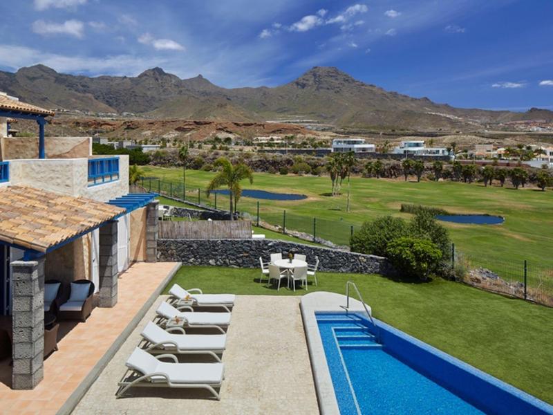 Moderner Pool mit Liegestühlen, Terrasse, Golfplatz und Bergen im Hintergrund bei blauem Himmel.