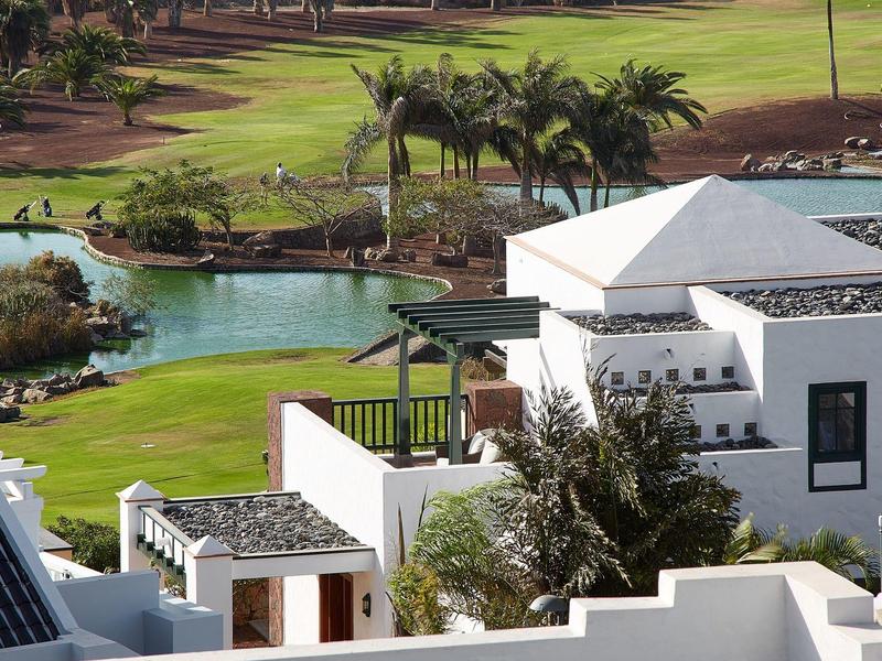 Weiße Häuser mit quadratischem Design liegen neben grünen Palmen und einem Teich mit blauem Wasser.