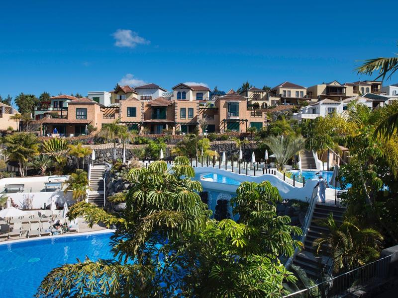 Große blaue Poolanlage mit Liegestühlen und Palmen vor Reihenhäusern und blauem Himmel.