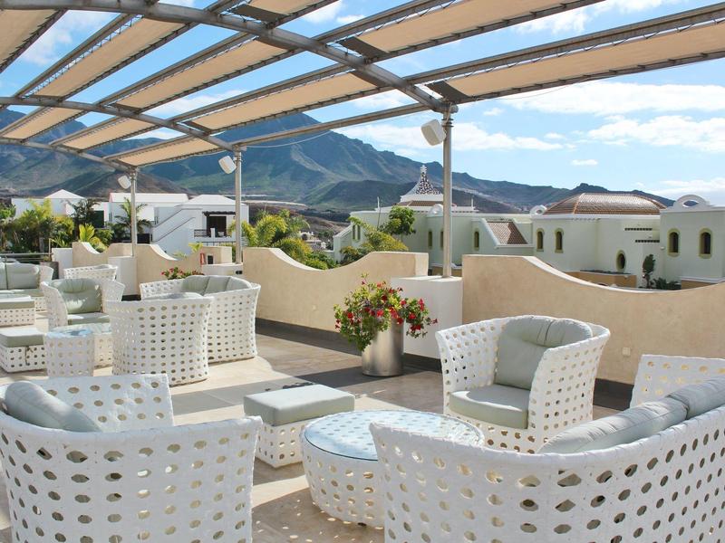 Dachterrasse mit weißem Kunststoffmöbel-Set, Glasdach, Pflanzen und Bergpanorama im Hintergrund.