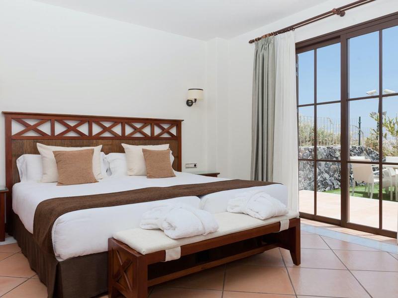 Gemütliches Schlafzimmer mit Holzmöbeln, großen Fenstern und hellem Fliesenboden.