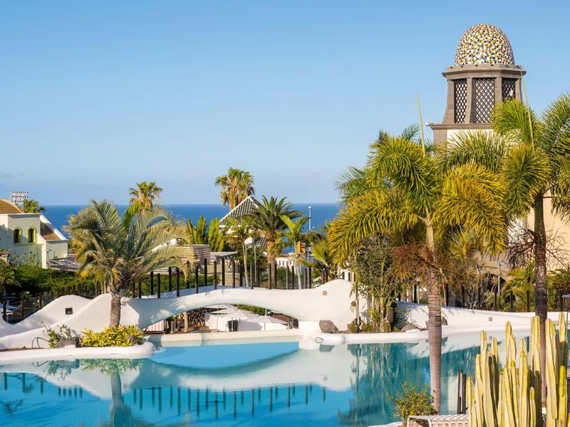 Blick auf einen Pool mit Brücke, Palmen und einem Turm vor klarem blauem Himmel und Meer.