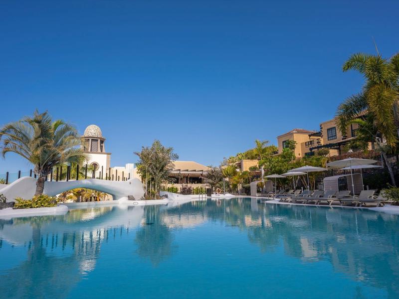 Großer blauer Pool vor weißen Gebäuden mit Palmen unter klarem blauem Himmel.