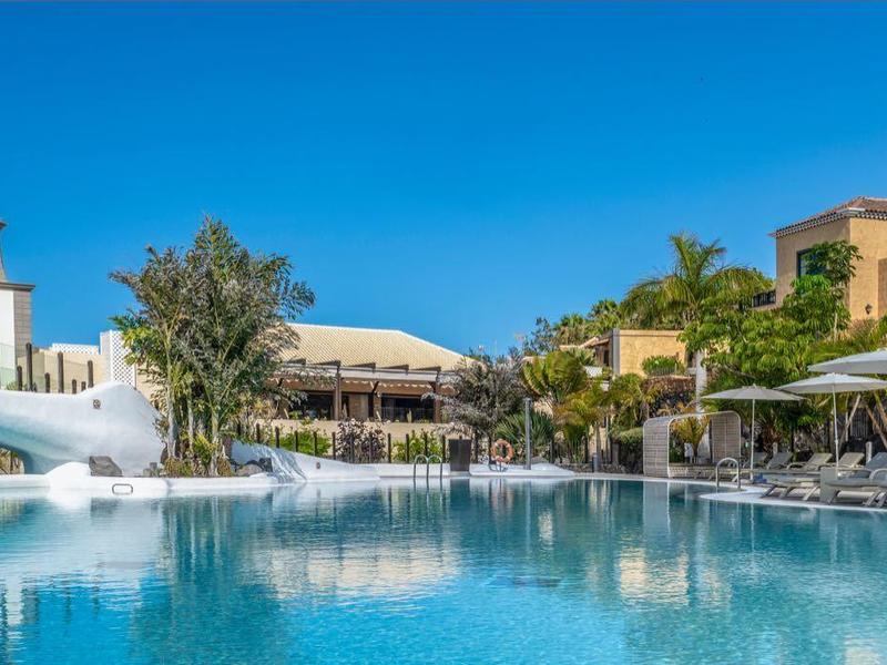 Hotelanlage mit großem Pool, Palmen, Liegestühlen und mehrstöckigen Gebäuden unter blauem Himmel.