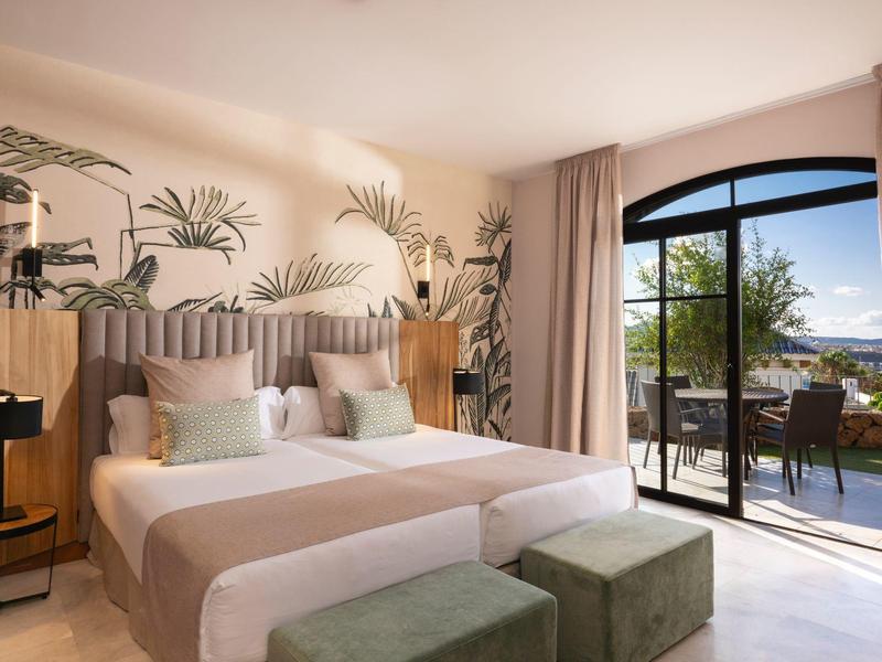 Helles Schlafzimmer mit zwei Betten, Pflanzenmuster an der Wand und Blick auf Terrasse mit Meer.