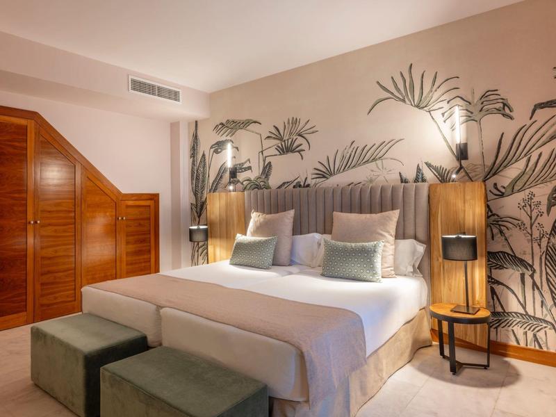 Helles Schlafzimmer mit großem Bett, gemusterter Wand, Holzschrank und zwei grünen Hockern.