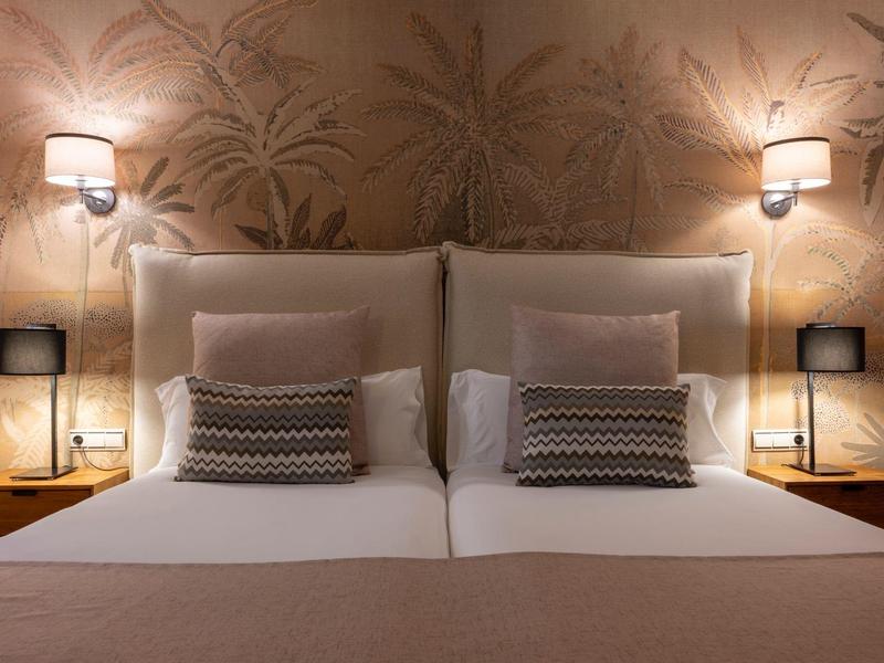 Symmetrisches Doppelbett mit beigen Kissen, Nachttischen, Lampen und dekorativer Tapete im Hintergrund.