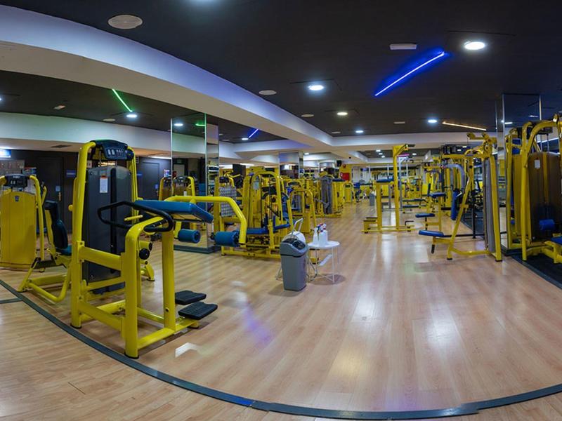 Großer Fitnessraum mit zahlreichen gelben Trainingsgeräten und hellem Holzfußboden.