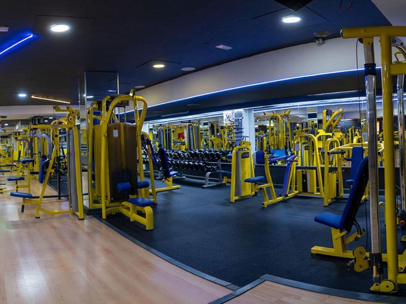 Großer Fitnessraum mit gelben Kraftgeräten und Hanteln, dunklem Boden und Holzparkett, helle Beleuchtung.