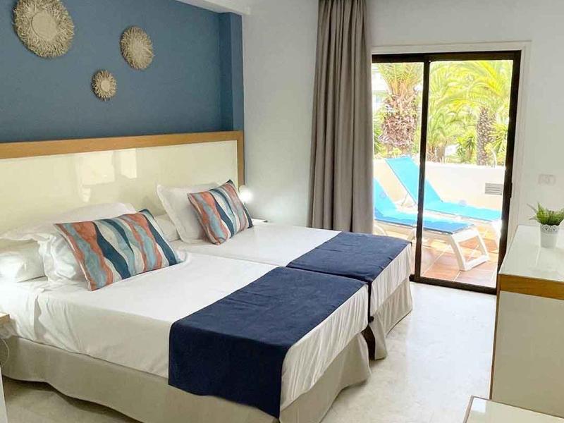 Helles Schlafzimmer mit zwei Einzelbetten, blauer Wand, Fenster zur Terrasse und moderner Einrichtung.