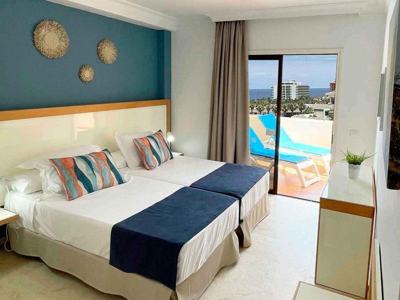 Modernes Hotelzimmer mit zwei Betten, bunten Kissen, Balkon und Blick auf den Pool.