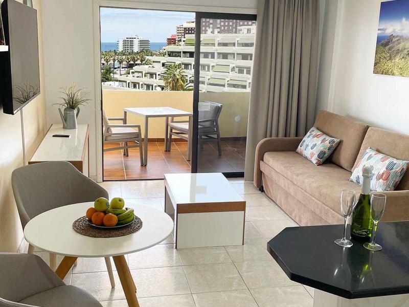 Helles Wohnzimmer mit Sofa, kleinem Esstisch, TV, Balkon und Panoramafenster.