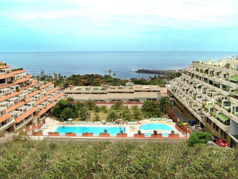 Zwei terrassenförmige Gebäude mit Schwimmbad in der Mitte, dahinter Meer und blauer Himmel.