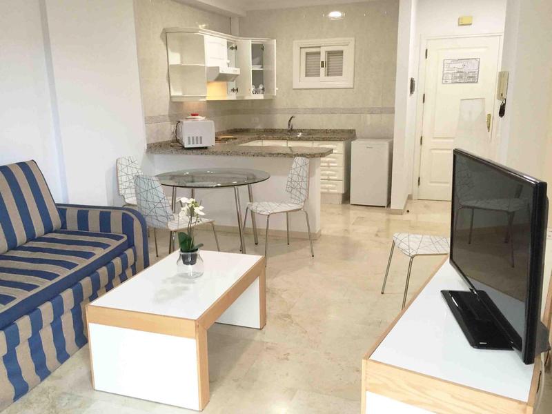 Kleines Wohnzimmer mit blau gestreiftem Sofa, weißem Couchtisch, Esstisch und Küche im Hintergrund.