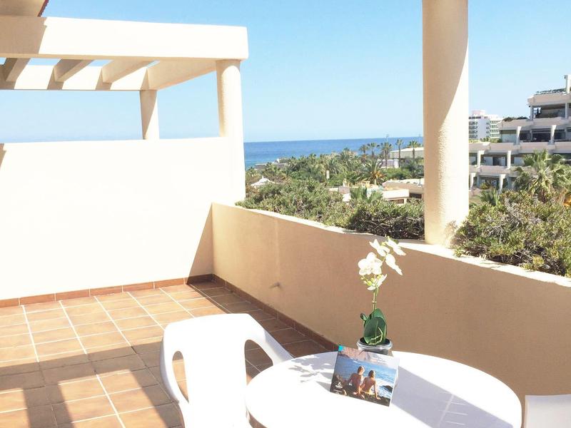 Balkon mit weißem Tisch und Stühlen, blauer Himmel und Meer im Hintergrund, helle Sonnenstrahlen.