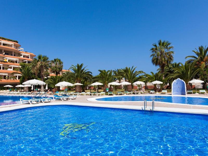 Großer blauer Pool mit Wasserfontänen, Sonnenschirmen und Palmen vor einem Hotel bei klarem Himmel.