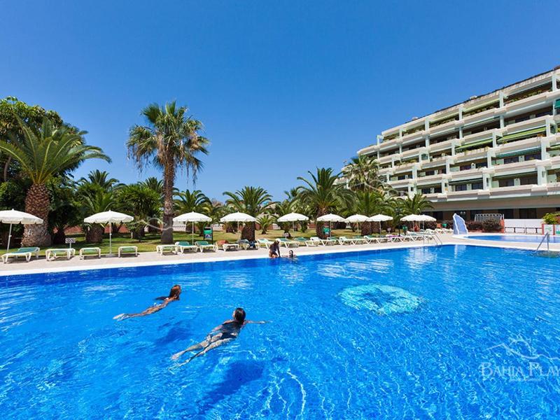 Ein großes Hotel mit Palmen und blauer Poollandschaft, darin schwimmen mehrere Personen.
