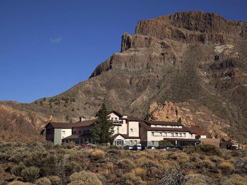 A lodge built at the base of a rugged, rocky mountain under a clear blue sky.