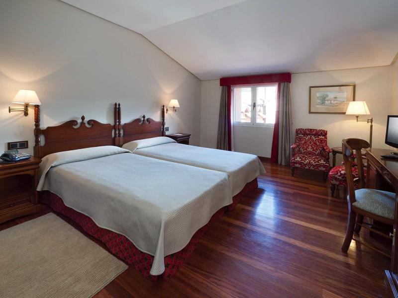 Cozy hotel room with two single beds, wooden flooring, red curtains, and classic furniture.