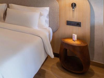 Nowoczesny pokój hotelowy z białym łóżkiem, drewnianą podłogą i okrągłą ciemną szafką nocną.