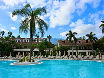 Großer Außenpool mit Palmen und Liegestühlen vor einem Hotel unter blauem Himmel.
