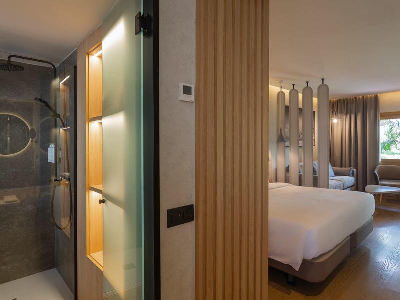 Nowoczesny pokój hotelowy z dużym łóżkiem, częścią wypoczynkową i nowoczesnym prysznicem z przeszklonymi drzwiami.