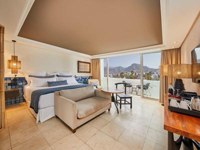 Habitación de hotel moderna con cama doble, área de estar y gran balcón con vista a la montaña.