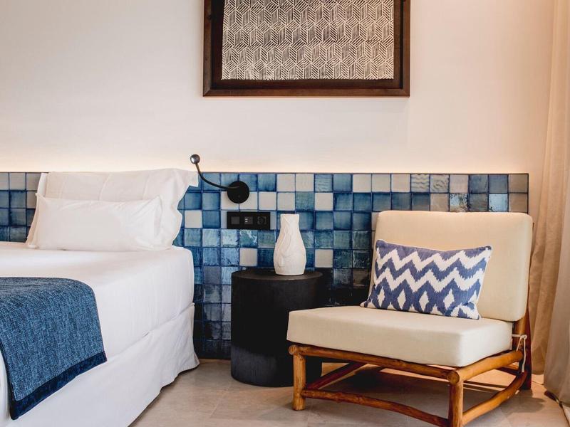 Habitación de hotel acogedora con cama blanca, acentos azules y un sillón con cojín estampado.