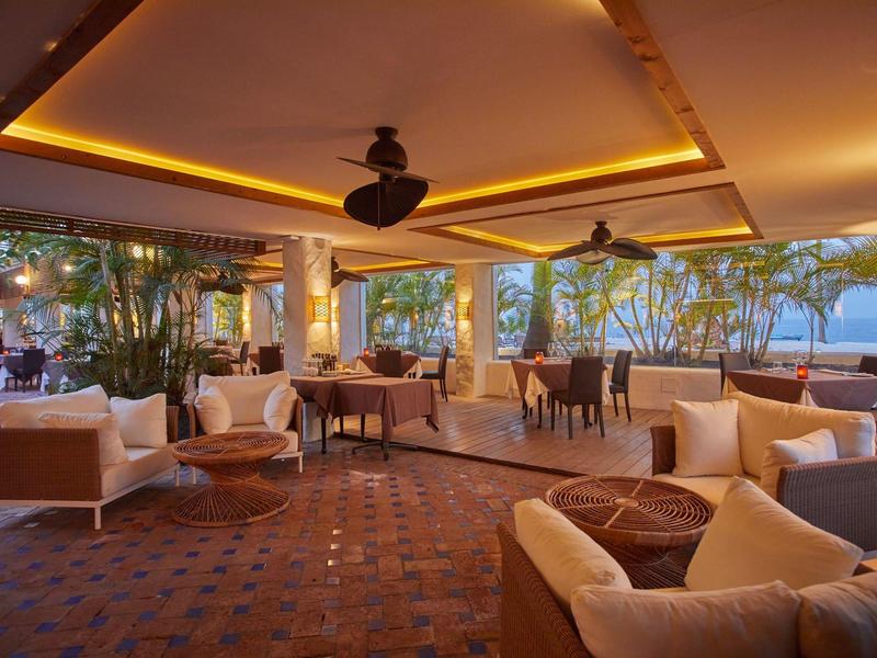 Terraza cubierta con áreas de asientos y ventiladores, rodeada de árboles con vista al agua.