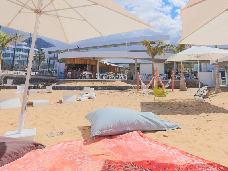 Plage de sable avec parasols, coussins et bar moderne en arrière-plan sous un ciel bleu.