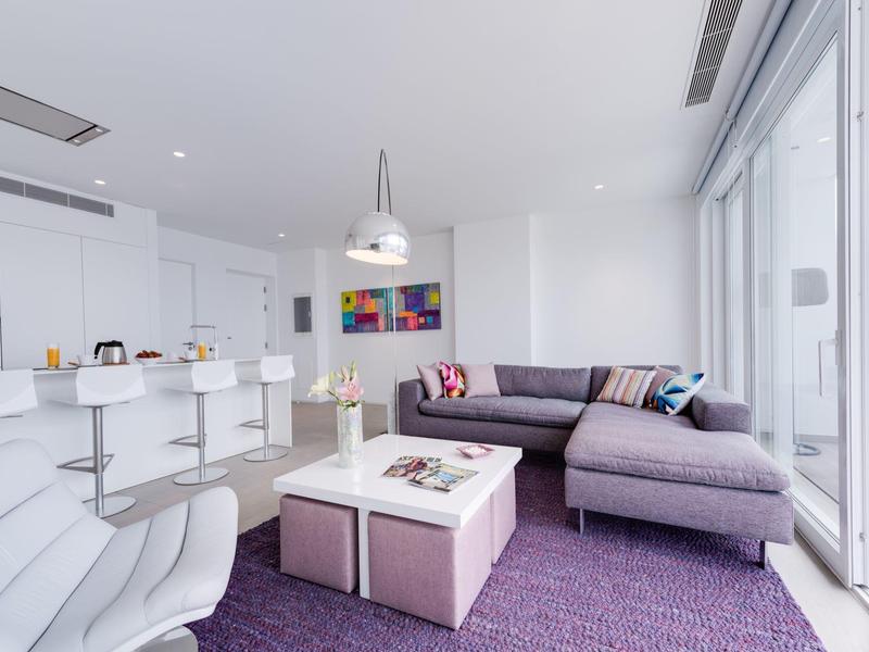 Espace de vie moderne avec tapis violet, canapé d'angle et cuisine attenante dans un appartement lumineux.