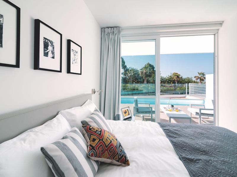 Chambre d'hôtel moderne avec lit, oreillers et vue sur la piscine à travers une baie vitrée.